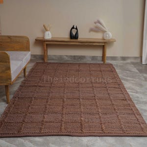 May include: A rectangular brown area rug featuring a raised grid pattern across its surface. The rug is positioned on a light-colored floor in front of a wooden bench and a wooden sofa frame. The text Theindoorrugs is visible on the rug surface.