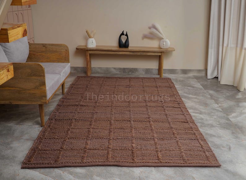 May include: A rectangular brown area rug featuring a raised grid pattern across its surface. The rug is positioned on a light-colored floor in front of a wooden bench and a wooden sofa frame. The text Theindoorrugs is visible on the rug surface.