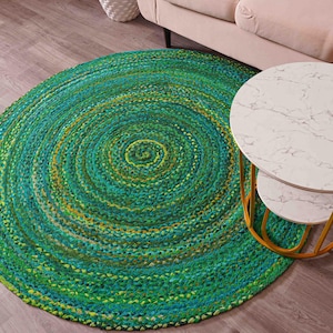 K&ouml;nnte beinhalten: Ein runder, geflochtener Teppich in Gr&uuml;n-, T&uuml;rkis- und Gelbt&ouml;nen. Der Teppich hat ein Spiralmuster und liegt auf einem Holzboden. Ein wei&szlig;er Beistelltisch mit Marmorplatte und goldenem Rahmen steht neben dem Teppich.