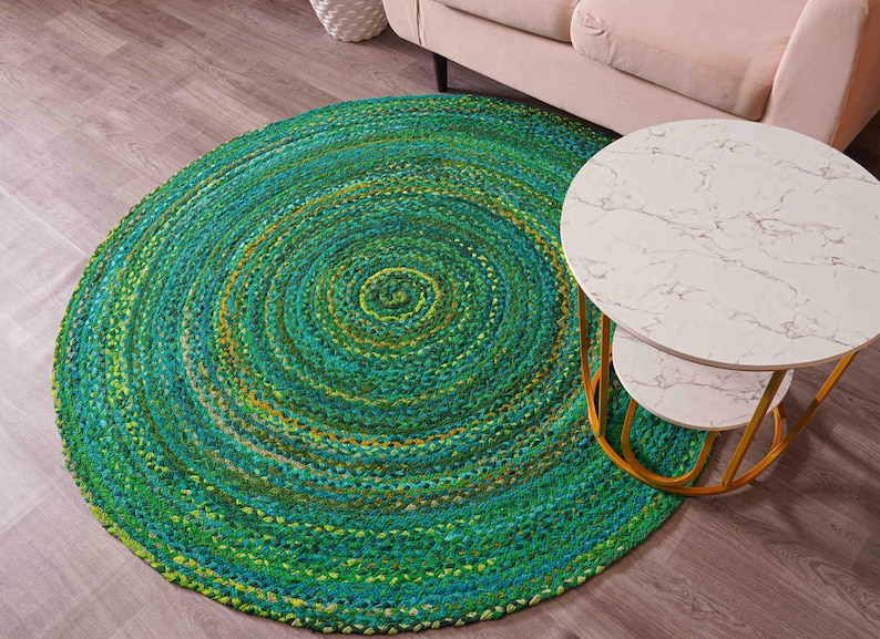 K&ouml;nnte beinhalten: Ein runder, geflochtener Teppich in Gr&uuml;n-, T&uuml;rkis- und Gelbt&ouml;nen. Der Teppich hat ein Spiralmuster und liegt auf einem Holzboden. Ein wei&szlig;er Beistelltisch mit Marmorplatte und goldenem Rahmen steht neben dem Teppich.