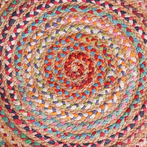 K&ouml;nnte beinhalten: Ein runder Jute-Teppich mit Spiralmuster. Der Teppich hat eine nat&uuml;rliche beige Farbe mit eingewebten Abschnitten aus rotem, blauem, gr&uuml;nem und wei&szlig;em Stoff. Das Design erzeugt ein strukturiertes, farbenfrohes Muster f&uuml;r die Inneneinrichtung.