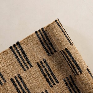 May include: Close-up of a woven rug with a natural beige background and black vertical stripe patterns. The rug has a textured surface and a rolled edge, showcasing a rustic aesthetic.