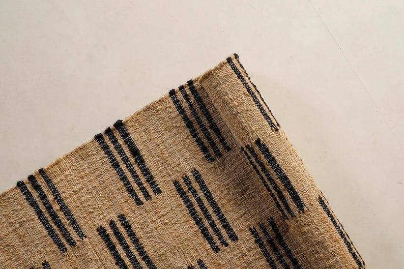 May include: Close-up of a woven rug with a natural beige background and black vertical stripe patterns. The rug has a textured surface and a rolled edge, showcasing a rustic aesthetic.
