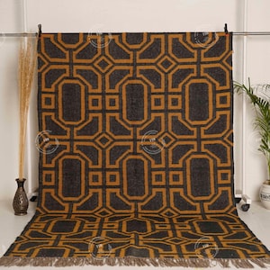 May include: A large, rectangular rug with a geometric pattern in shades of charcoal gray and gold. The design features interlocking shapes and a fringed edge. The rug is displayed on a white background.