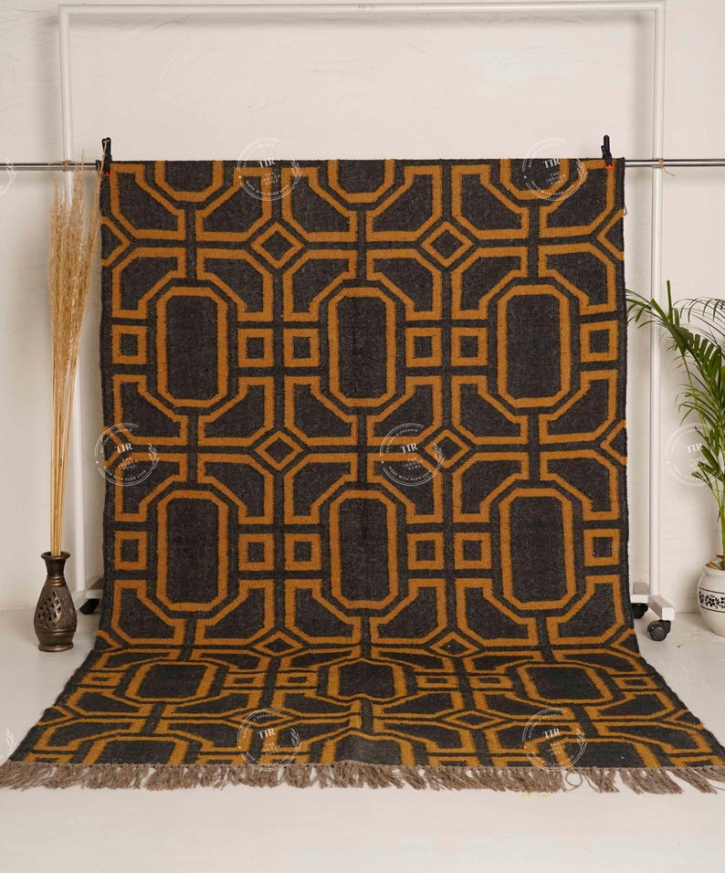 May include: A large, rectangular rug with a geometric pattern in shades of charcoal gray and gold. The design features interlocking shapes and a fringed edge. The rug is displayed on a white background.