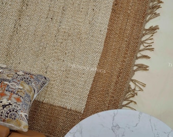 Puede incluir: Una alfombra rectangular de yute con un centro beige natural y un borde marrón. La alfombra tiene un borde con flecos y se combina con un cojín estampado y una mesa con tapa de mármol. El texto "THE INDOOR RUGS" es visible en la alfombra.