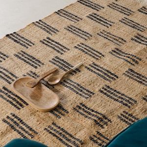 May include: A woven jute rug with a geometric pattern of black stripes. A wooden serving tray and two wooden spoons rest on the rug. The rug has a natural, neutral color and is placed near a teal-colored sofa.