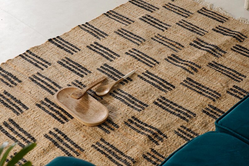 May include: A woven jute rug with a geometric pattern of black stripes. A wooden serving tray and two wooden spoons rest on the rug. The rug has a natural, neutral color and is placed near a teal-colored sofa.