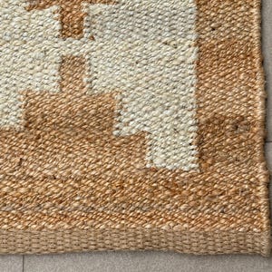 May include: A woven area rug featuring a natural tan jute border and a central section of off-white fibres. The design displays a geometric pattern with stepped, block-like shapes in tan jute that contrast against the lighter off-white background.