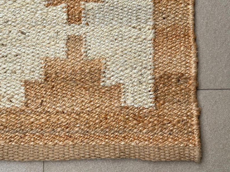 May include: A woven area rug featuring a natural tan jute border and a central section of off-white fibres. The design displays a geometric pattern with stepped, block-like shapes in tan jute that contrast against the lighter off-white background.