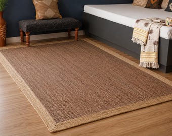 May include: A rectangular area rug featuring a dark brown woven center panel surrounded by a wide, light tan braided border. The rug is constructed from natural fibers with a visible textured weave, creating a distinct contrast between the dark interior and the lighter, thicker outer edge.