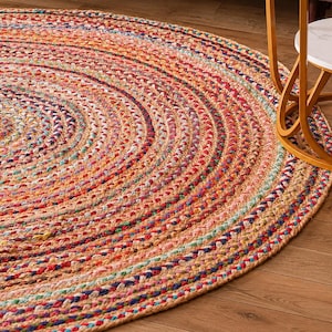Può includere: Un tappeto rotondo caratterizzato da una struttura intrecciata realizzata con strisce di tessuto multicolore. Il design è composto da cerchi concentrici nei toni dell'arancione, rosso, blu, giallo e marrone chiaro, creando una superficie tessuta.