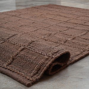 May include: A brown area rug featuring a raised grid pattern of squares across its surface. The rug is constructed with a thick, textured weave and is partially rolled at one corner, revealing the depth and density of the material construction.