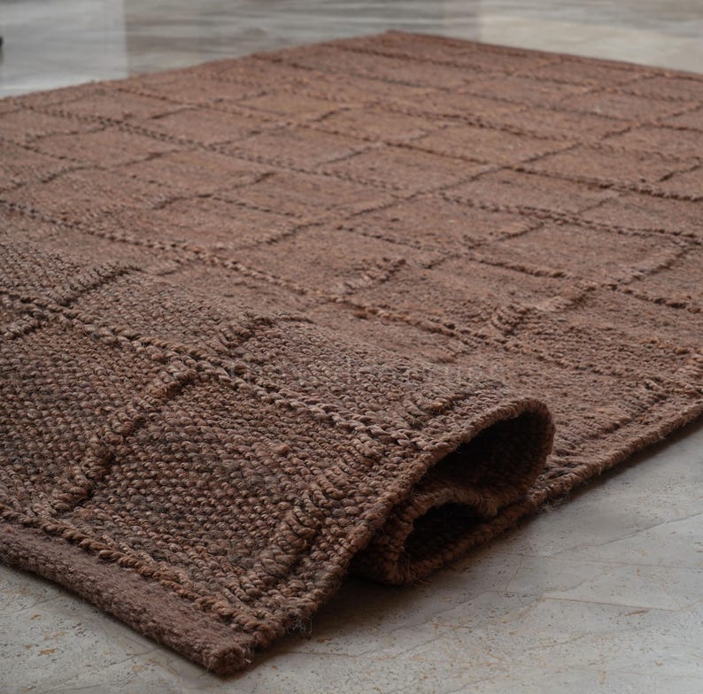 May include: A brown area rug featuring a raised grid pattern of squares across its surface. The rug is constructed with a thick, textured weave and is partially rolled at one corner, revealing the depth and density of the material construction.