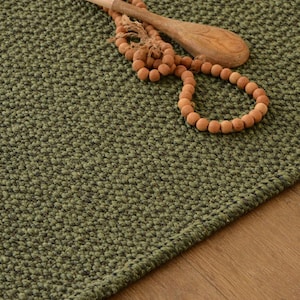 May include: A green textured rug with a wooden spoon and a beaded necklace. The rug has a woven appearance and is a muted green color. The wooden spoon is light brown, and the necklace has round, light brown beads.