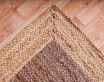 May include: A corner of a braided jute rug in shades of tan and brown. The rug's texture is visible, with a lighter border and a darker center. The rug is placed on a wooden floor with a herringbone pattern.