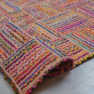 May include: A rectangular area rug featuring a woven pattern of natural tan jute fibers interspersed with vibrant, multi-colored fabric strips in shades of pink, blue, yellow, green, and orange, arranged in a repeating grid of alternating blocks.