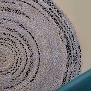 Puede incluir: Una alfombra redonda trenzada con un patrón concéntrico de fibras tejidas en tonos blanco, gris claro y negro. La textura se crea mediante gruesos hilos trenzados dispuestos en una formación circular que irradia desde el centro al borde.