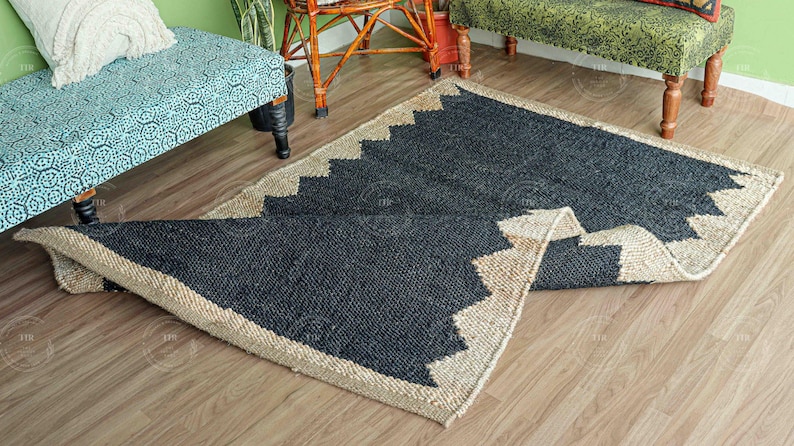 Op de afbeelding: Een rechthoekig jute vloerkleed met een zwart midden en een natuurlijke beige rand. De rand heeft een zigzagpatroon. Het vloerkleed is gedeeltelijk opgerold, waardoor de onderkant zichtbaar is. Het vloerkleed ligt op een lichte houten vloer.