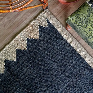 Op de afbeelding: Een hoek van een jute vloerkleed met een geometrisch ontwerp. Het vloerkleed heeft een donkerblauw midden met een beige rand en zigzagpatroon. Een groen kussen met patroon en een houten stoel staan op de achtergrond.