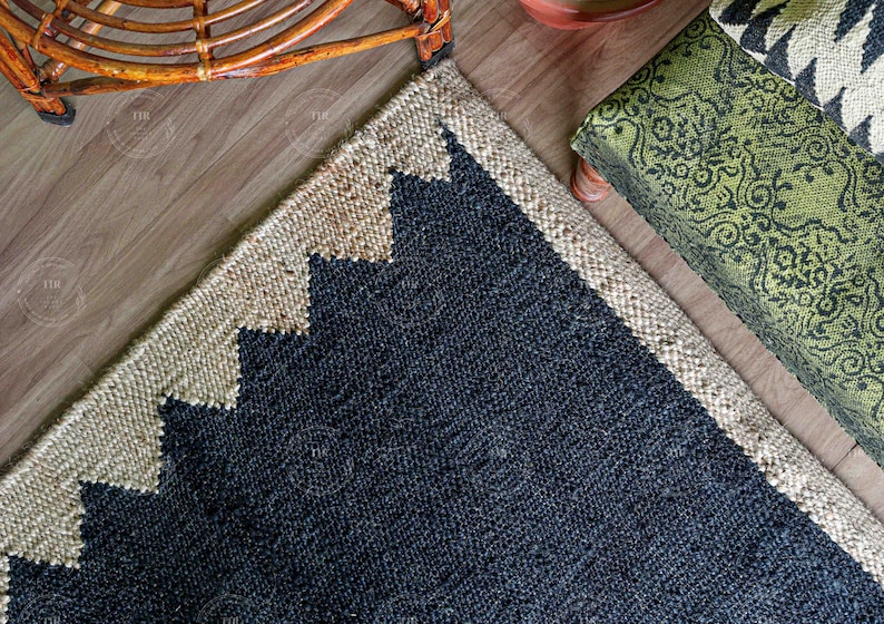 Op de afbeelding: Een hoek van een jute vloerkleed met een geometrisch ontwerp. Het vloerkleed heeft een donkerblauw midden met een beige rand en zigzagpatroon. Een groen kussen met patroon en een houten stoel staan op de achtergrond.