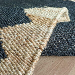 Op de afbeelding: Close-up van een geweven jute vloerkleed met een geometrisch ontwerp. Het vloerkleed heeft een combinatie van donkerblauwe en natuurlijke beige tinten. De textuur van het vloerkleed is zichtbaar en toont de natuurlijke vezels en het ingewikkelde weefpatroon. Het vloerkleed is gedeeltelijk opgerold.