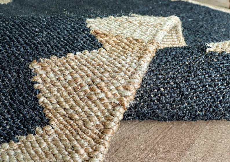 Op de afbeelding: Close-up van een geweven jute vloerkleed met een geometrisch ontwerp. Het vloerkleed heeft een combinatie van donkerblauwe en natuurlijke beige tinten. De textuur van het vloerkleed is zichtbaar en toont de natuurlijke vezels en het ingewikkelde weefpatroon. Het vloerkleed is gedeeltelijk opgerold.