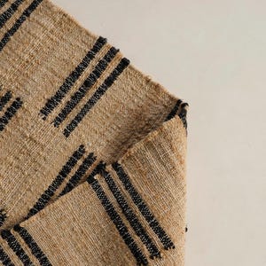 May include: A tan woven rug with black stripes arranged in a diagonal pattern. The rug is folded over, revealing the underside. The texture of the rug is visible, and the background is a plain, off-white color.