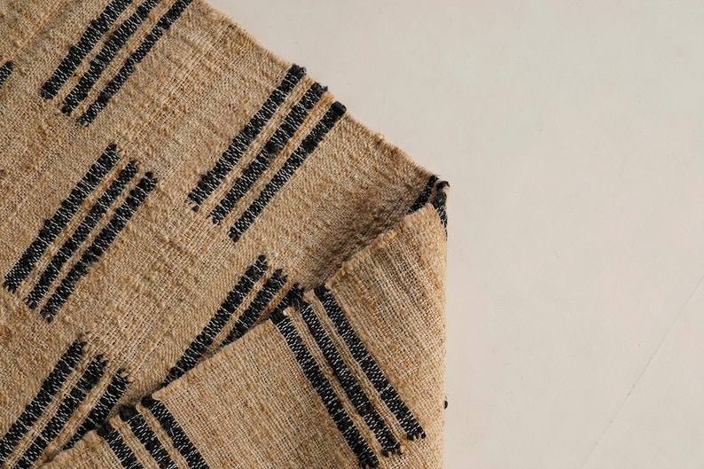 May include: A tan woven rug with black stripes arranged in a diagonal pattern. The rug is folded over, revealing the underside. The texture of the rug is visible, and the background is a plain, off-white color.