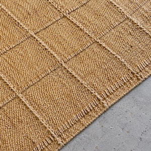 May include: A golden-tan woven rug featuring a prominent grid pattern created by raised, textured lines. The surface displays a dense, uniform weave with a consistent color throughout, forming a series of square sections across the material.