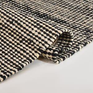 May include: A woven area rug featuring a high-contrast pattern of alternating black and natural beige fibres. The rug is folded over at one corner, revealing the thick, textured weave and the dense, durable construction of the material.