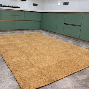 May include: A rectangular, woven jute rug with a geometric square pattern. The rug is a warm, natural tan color and lies on a gray concrete floor. The rug is in a kitchen setting with green cabinets.