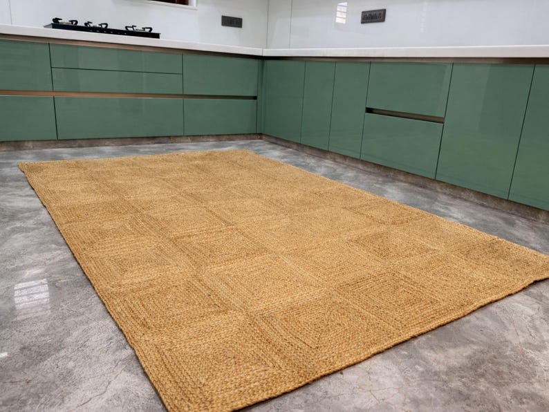 May include: A rectangular, woven jute rug with a geometric square pattern. The rug is a warm, natural tan color and lies on a gray concrete floor. The rug is in a kitchen setting with green cabinets.