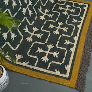 Könnte beinhalten: Ein dunkelblauer Teppich mit einem geometrischen Muster in Off-White. Der Teppich hat einen senfgelben Rand und graue Fransen. Eine Topfpflanze mit grünem Laub und ein dekorativer Topf mit Steinen befinden sich im Vordergrund.