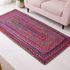 Puede incluir: Una alfombra rectangular trenzada con una vibrante mezcla de colores, incluyendo rosa, morado, azul y amarillo. La alfombra tiene un borde definido y está colocada sobre un suelo de color claro. Es un artículo de decoración del hogar.