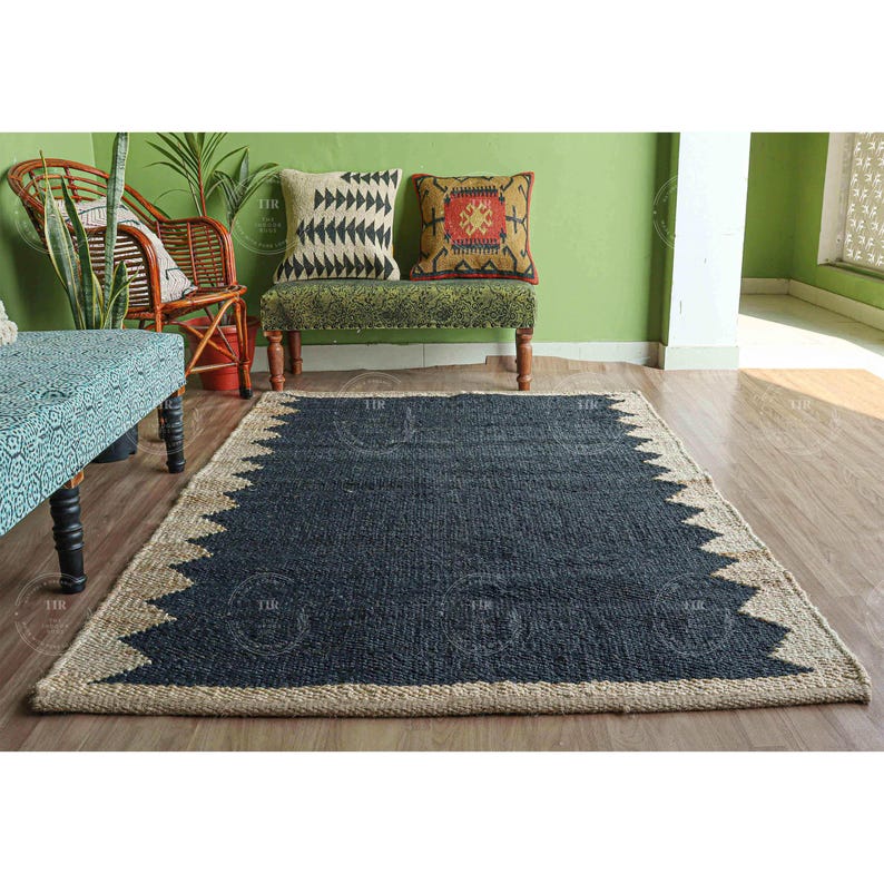 Op de afbeelding: Een rechthoekig, marineblauw jute vloerkleed met een zigzagrand in een natuurlijke, beige kleur. Het kleed ligt op een houten vloer. De achtergrond toont een groene muur, een bank met patronen en een rieten stoel.