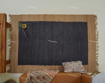 May include: A rectangular jute rug with a dark gray center and a natural tan border. The rug has a fringed edge and is displayed on a wall. A decorative pillow and throw are on a brown leather couch below the rug. A small yellow decorative object is attached to the rug.