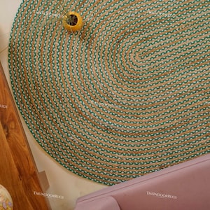 Puede incluir: Alfombra ovalada de yute con un dise&ntilde;o trenzado verde y beige. La alfombra est&aacute; colocada sobre un suelo claro, con una mesa de madera y un sof&aacute; rosa en el fondo. Un peque&ntilde;o jarr&oacute;n amarillo con una planta est&aacute; sobre la alfombra.