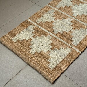 May include: A rectangular woven runner rug featuring a natural tan jute base with a repeating geometric pattern in off-white. The design consists of symmetrical, stepped diamond shapes arranged in a linear sequence across the textured surface.
