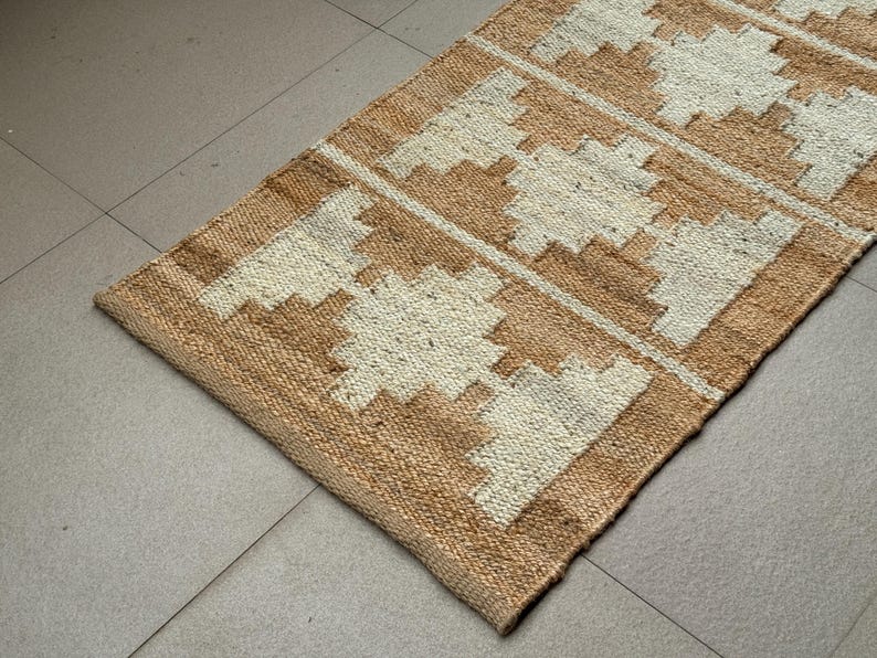 May include: A rectangular woven runner rug featuring a natural tan jute base with a repeating geometric pattern in off-white. The design consists of symmetrical, stepped diamond shapes arranged in a linear sequence across the textured surface.