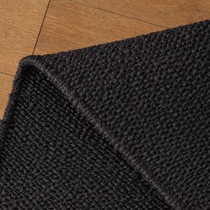 May include: A close-up of a section of a black woven rug. The rug has a textured surface and a folded corner, revealing the thickness of the material. The rug is placed on a wooden surface.
