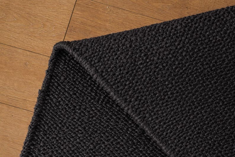 May include: A close-up of a section of a black woven rug. The rug has a textured surface and a folded corner, revealing the thickness of the material. The rug is placed on a wooden surface.