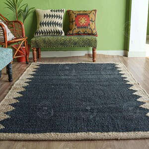 Op de afbeelding: Een rechthoekig jute vloerkleed met een donkerblauw midden en een natuurlijke beige rand. De rand heeft een zigzagpatroon. Het vloerkleed ligt op een houten vloer, met meubels en planten op de achtergrond.