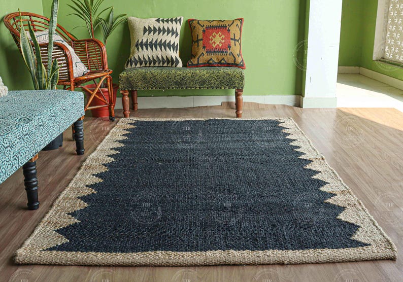 Op de afbeelding: Een rechthoekig jute vloerkleed met een donkerblauw midden en een natuurlijke beige rand. De rand heeft een zigzagpatroon. Het vloerkleed ligt op een houten vloer, met meubels en planten op de achtergrond.