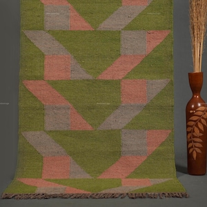 May include: A rectangular woven rug featuring a repeating geometric pattern in olive green, muted pink, and grey. The rug has a flat-weave texture and a short fringe along the bottom edge. The design consists of interlocking shapes.