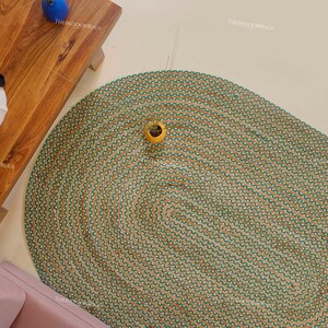 Puede incluir: Una alfombra ovalada con base de yute natural y borde verde. La alfombra est&aacute; colocada sobre un suelo de color claro. Una mesa de madera y un sof&aacute; rosa son parcialmente visibles. La alfombra es un art&iacute;culo de decoraci&oacute;n del hogar.