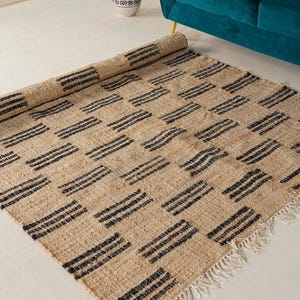 May include: A rectangular beige rug with a repeating pattern of black horizontal stripes arranged in a grid. The rug is partially rolled up, revealing its woven texture and fringed edges. The rug is placed on a white floor.