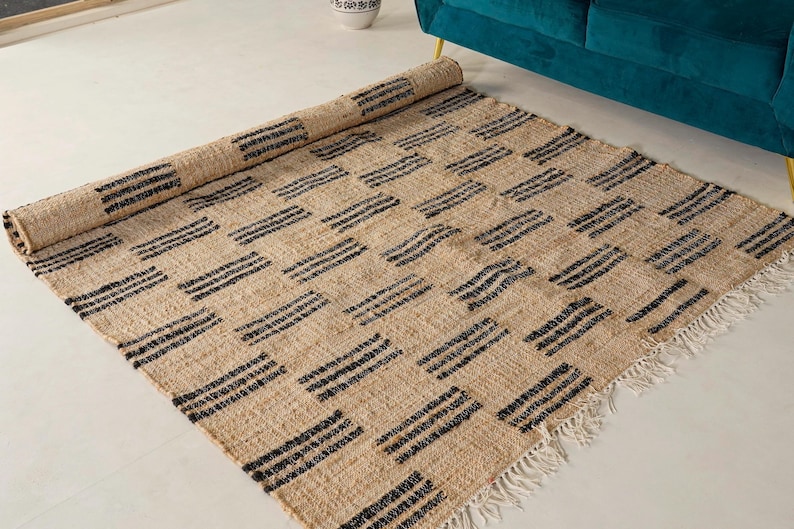 May include: A rectangular beige rug with a repeating pattern of black horizontal stripes arranged in a grid. The rug is partially rolled up, revealing its woven texture and fringed edges. The rug is placed on a white floor.