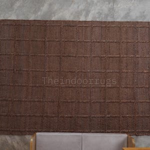 May include: A rectangular brown rug featuring a woven grid pattern of raised squares. The surface has a textured, natural fiber appearance. The text "Theindoorrugs" is printed in a light color across the center of the rug.