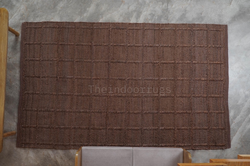 May include: A rectangular brown rug featuring a woven grid pattern of raised squares. The surface has a textured, natural fiber appearance. The text "Theindoorrugs" is printed in a light color across the center of the rug.